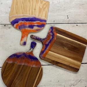 May include: Three wooden cutting boards with handles. Two are rectangular, and one is round. Each board has a unique design with a resin finish in shades of orange, purple, and white. The boards are on a white wooden surface.