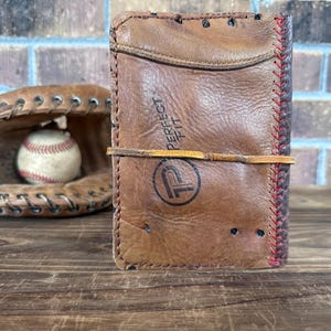 K&ouml;nnte beinhalten: Ein braunes Lederportemonnaie mit dem Text "PERFECT FIT" und einem Logo, gesichert mit einem Lederband. Ein Baseballhandschuh und ein Baseball befinden sich im Hintergrund. Das Portemonnaie scheint aus recyceltem Baseballhandschuhleder zu bestehen.