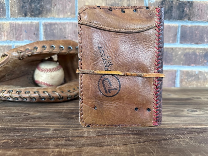 K&ouml;nnte beinhalten: Ein braunes Lederportemonnaie mit dem Text "PERFECT FIT" und einem Logo, gesichert mit einem Lederband. Ein Baseballhandschuh und ein Baseball befinden sich im Hintergrund. Das Portemonnaie scheint aus recyceltem Baseballhandschuhleder zu bestehen.