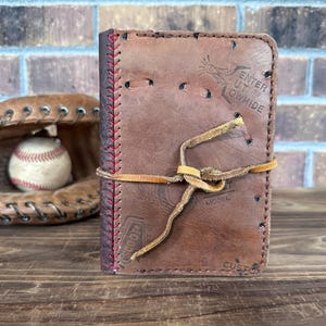 K&ouml;nnte beinhalten: Ein braunes Leder-Notizbuch mit roten N&auml;hten und einem Lederbandverschluss. Das Notizbuch besteht aus recyceltem Baseballmaterial und enth&auml;lt Text vom urspr&uuml;nglichen Baseball. Ein Baseball und ein Handschuh sind im Hintergrund.