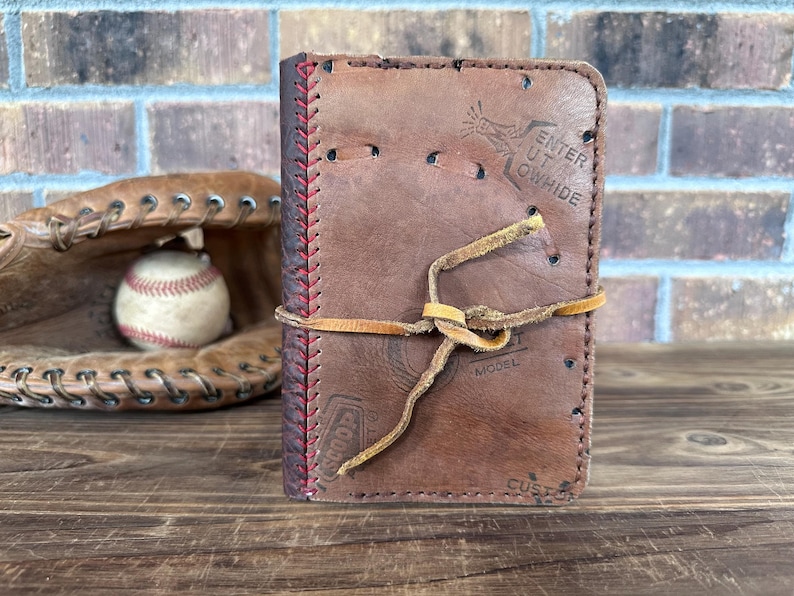 K&ouml;nnte beinhalten: Ein braunes Leder-Notizbuch mit roten N&auml;hten und einem Lederbandverschluss. Das Notizbuch besteht aus recyceltem Baseballmaterial und enth&auml;lt Text vom urspr&uuml;nglichen Baseball. Ein Baseball und ein Handschuh sind im Hintergrund.