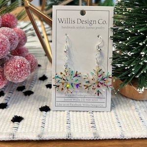 May include: A pair of silver snowflake earrings with iridescent glitter. The earrings are on a white card with the text "Willis Design Co. handmade. stylish. leather. jewelry shop online at: www.willisdesignco.com Jeanne Willis 231-499-9029 Like us on Facebook: Willis Design Co."