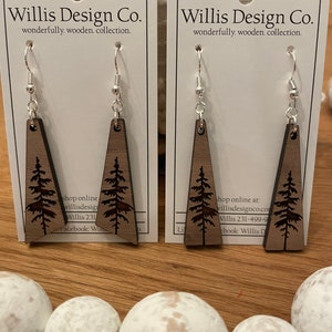May include: A pair of wooden earrings with a tree design. The earrings are brown and have silver hooks. The earrings are on a white card with the text "Willis Design Co. wonderfully. wooden. collection." printed on it.