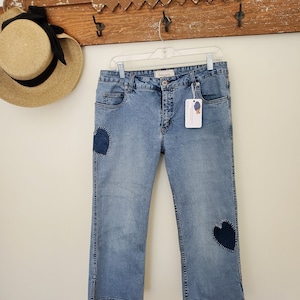 May include: Light blue denim jeans with dark blue heart-shaped patches on the front. The jeans are hanging on a wooden rack with a straw hat.