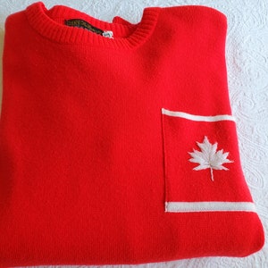 May include: Red sweater with a white pocket and a white embroidered maple leaf. The sweater has a ribbed crew neck.