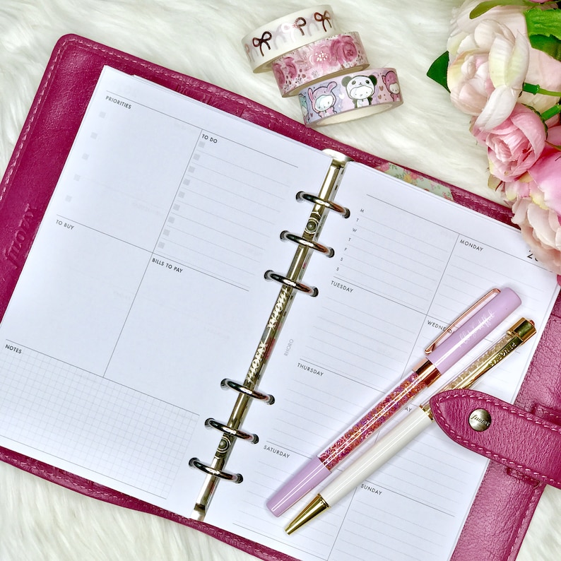 May include: A pink leather planner with a white weekly and daily planner insert. The planner has a gold ring binder and is open to show the weekly and daily pages. There are two sparkly pens on the planner, one pink and one white.