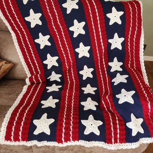 May include: A crocheted blanket with red, white, and blue stripes and white stars. The blanket is folded on a couch.