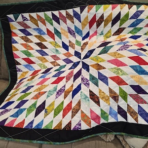 May include: A colorful quilt with a starburst design. The quilt features a central star pattern radiating outwards with diamond-shaped patches in various colors, including red, blue, green, and purple. It has a black border with a decorative stitch.
