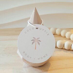 May include: A light beige, cone-shaped object with a white string holding a round tag. The tag features the text "COCO UTTIE BODY + HOME" and a palm tree illustration in a light brown colour. A wooden bead strand is visible in the background.