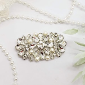 May include: A silver and crystal bridal brooch with a cluster of faux pearls. The brooch is surrounded by white lace and green leaves.