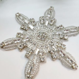 May include: A silver snowflake-shaped embellishment with crystal rhinestones and beads. The snowflake is made of a delicate, intricate design.