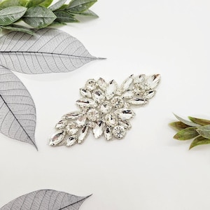 May include: A sparkling silver and clear crystal embellishment in a floral design. The embellishment features various shapes and sizes of crystals. The background is white with green and grey leaves.