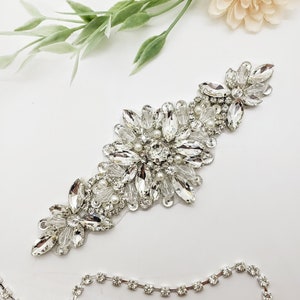 May include: A sparkling, silver-toned crystal and pearl embellishment. The design features a central cluster of large, faceted crystals surrounded by smaller stones and delicate pearls. The piece is elongated and floral-inspired, perfect for bridal or formal wear.