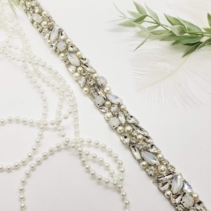 Pode incluir: Uma fita de contas prateadas com cristais e pérolas transparentes e brancas. A fita está sobre um fundo branco com um fio de pérolas brancas.