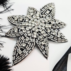 Puede incluir: Un adorno en forma de estrella con base negra, adornado con cuentas plateadas y brillantes pedrería. También hay plumas negras y una cinta de satén negra, creando un diseño glamuroso. Ideal para añadir brillo a cualquier atuendo.