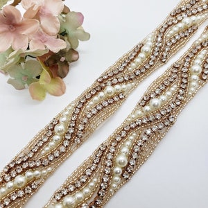 May include: Two decorative beaded trims with a wave pattern. The trim features alternating rows of small gold beads, clear rhinestones, and large ivory pearls. The background is white, and a small bouquet of pink and green flowers is in the upper left corner.