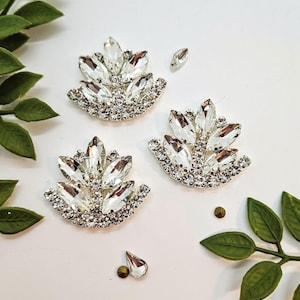 Peut inclure: Trois ornements décoratifs en forme de feuilles ou d'éventails, composés de strass transparents taille marquise et de petits cristaux ronds sertis sur une base en métal argenté, accompagnés de plusieurs cristaux individuels épars.