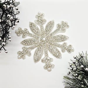 May include: A silver snowflake decoration with a beaded design and clear crystal accents. The snowflake has six points, each with a rounded tip and a pearl at the end. The centre of the snowflake has a larger pearl.