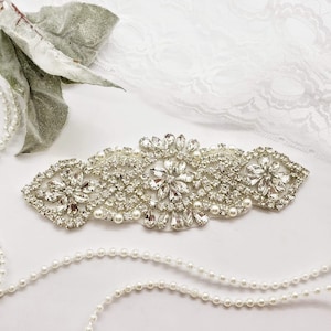 May include: A silver rhinestone and pearl embellishment with a floral design. The embellishment is on a white background with a pearl necklace.