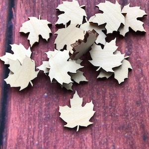May include: A pile of wooden maple leaf shapes, scattered on a dark red background. The leaves are unfinished and ready to be painted or decorated.