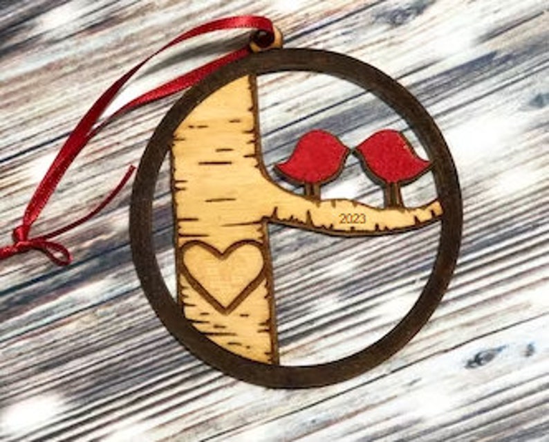 K&ouml;nnte beinhalten: Ein h&ouml;lzerner Weihnachtsbaumschmuck mit einem Birkenbaumdesign, zwei roten V&ouml;geln, die auf einem Ast sitzen, einem in den Baumstamm geschnitzten Herzen und dem Jahr 2023, das auf den Ast eingraviert ist.