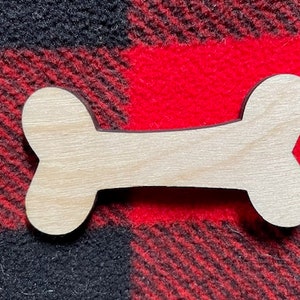 May include: A wooden dog bone cutout, unpainted, with a smooth surface. The bone is on a red and black plaid fabric background.