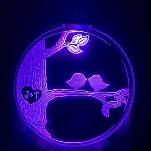 K&ouml;nnte beinhalten: Ein violett beleuchteter Holzkreis mit einem geschnitzten Baum, zwei V&ouml;geln und einem Herzen mit den Initialen "J+T".