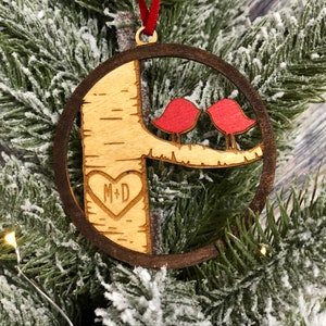 K&ouml;nnte beinhalten: Ein h&ouml;lzerner Weihnachtsbaumschmuck mit zwei roten V&ouml;geln, die auf einem Ast sitzen, und einem in den Baumstamm geschnitzten Herzen. Das Herz tr&auml;gt die Initialen "M+D" im Inneren.