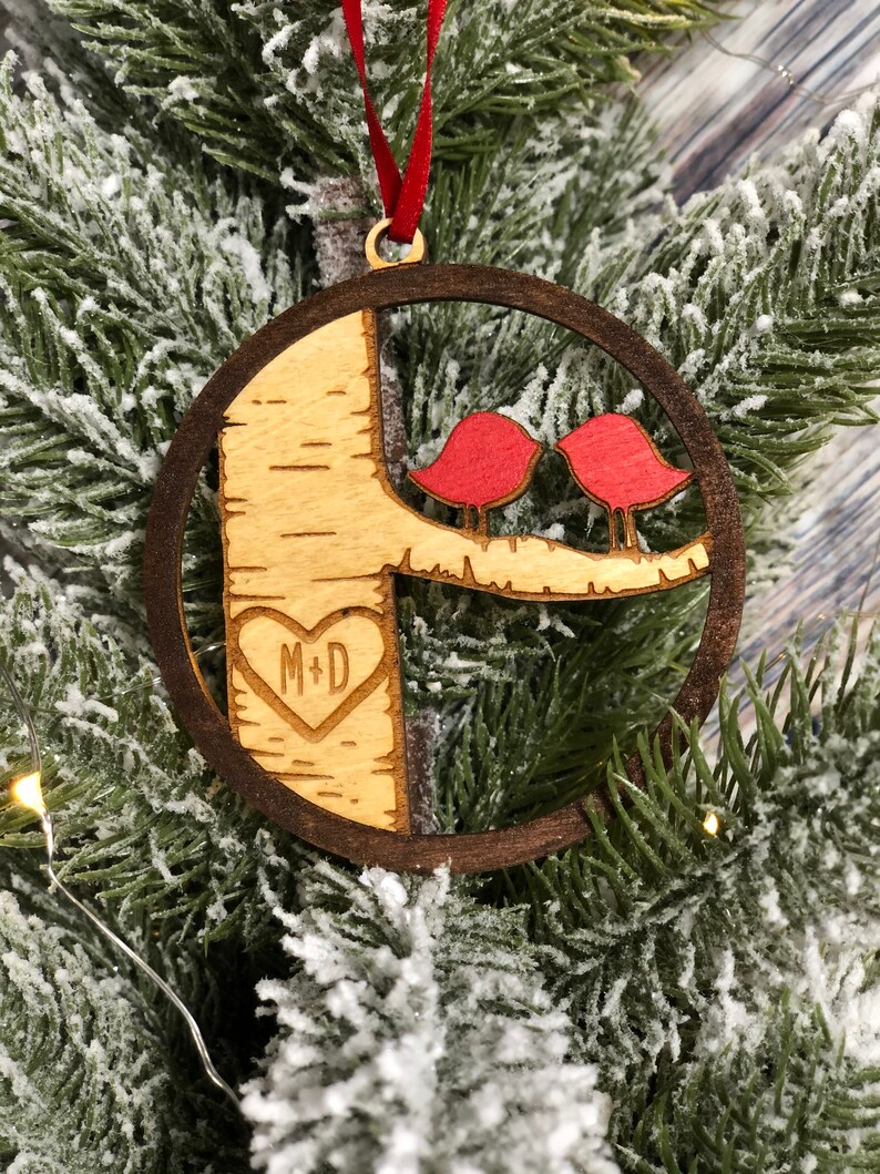 K&ouml;nnte beinhalten: Ein h&ouml;lzerner Weihnachtsbaumschmuck mit zwei roten V&ouml;geln, die auf einem Ast sitzen, und einem in den Baumstamm geschnitzten Herzen. Das Herz tr&auml;gt die Initialen "M+D" im Inneren.