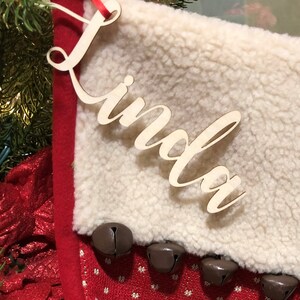 May include: A white faux fur stocking with a red cuff and jingle bells. The stocking has a wooden name tag with the name "Linda" written in cursive.