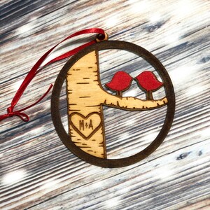 K&ouml;nnte beinhalten: Ein runder Weihnachtsbaumschmuck aus Holz mit einem Baum und zwei roten V&ouml;geln. Der Baum hat ein Herz mit den Initialen "M+A" eingegraben.