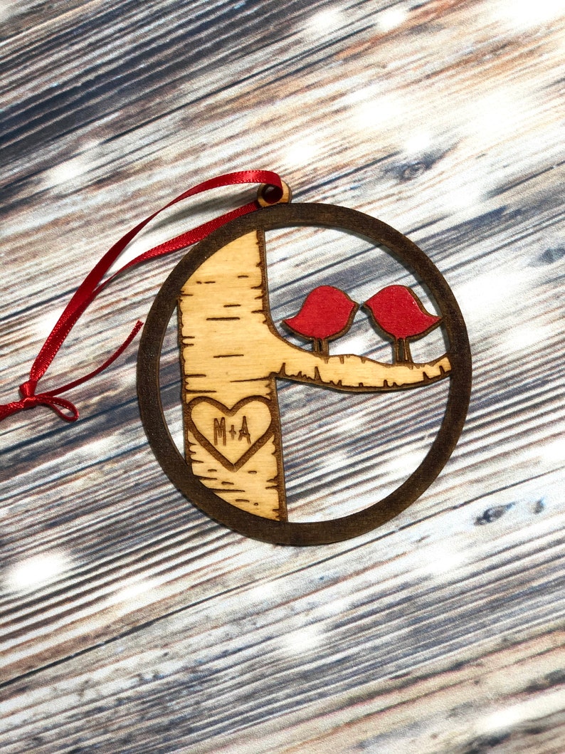 K&ouml;nnte beinhalten: Ein runder Weihnachtsbaumschmuck aus Holz mit einem Baum und zwei roten V&ouml;geln. Der Baum hat ein Herz mit den Initialen "M+A" eingegraben.