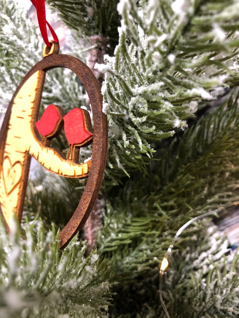 K&ouml;nnte beinhalten: Ein h&ouml;lzerner Weihnachtsbaumschmuck in Form eines Halbmondes mit zwei roten V&ouml;geln, die auf einem Ast sitzen. Der Schmuck h&auml;ngt an einem verschneiten k&uuml;nstlichen Weihnachtsbaum.