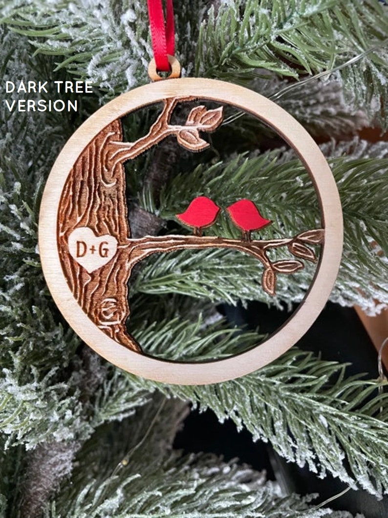 K&ouml;nnte beinhalten: Ein h&ouml;lzerner Weihnachtsbaumschmuck mit einem geschnitzten Baum, zwei roten V&ouml;geln und den Initialen "D+G" in einem Herzen. Der Schmuck h&auml;ngt an einem Weihnachtsbaum.