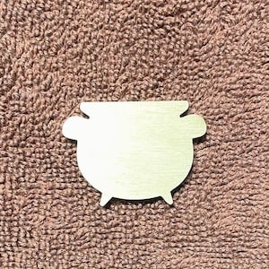 May include: A wooden cauldron-shaped cutout with three small legs and two side handles. The cutout is light-colored and sits on a brown, textured surface. This item is suitable for crafting or decorative purposes.