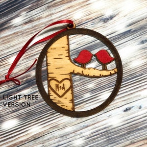 K&ouml;nnte beinhalten: Ein h&ouml;lzerner Weihnachtsbaumschmuck in Form eines Baumes mit zwei roten V&ouml;geln, die auf einem Ast sitzen. Der Baum hat ein Herz mit den Initialen "M+A" eingegraben. Der Schmuck h&auml;ngt an einem roten Band. "LIGHT TREE VERSION" steht unter dem Schmuck.