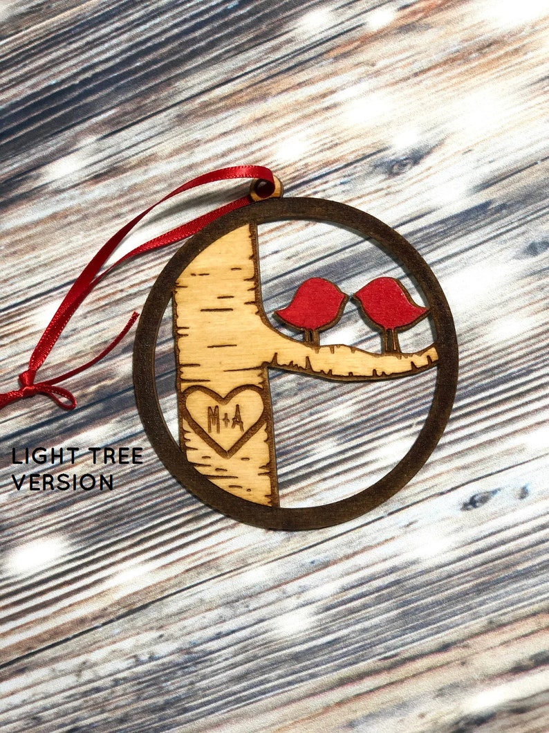 K&ouml;nnte beinhalten: Ein h&ouml;lzerner Weihnachtsbaumschmuck in Form eines Baumes mit zwei roten V&ouml;geln, die auf einem Ast sitzen. Der Baum hat ein Herz mit den Initialen "M+A" eingegraben. Der Schmuck h&auml;ngt an einem roten Band. "LIGHT TREE VERSION" steht unter dem Schmuck.