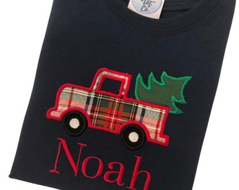 May include: A navy blue t-shirt featuring an embroidered applique of a plaid truck carrying a green Christmas tree. The truck is outlined in red thread, and the name Noah is embroidered in red below the truck. A Luxe Tot Co. tag is attached.