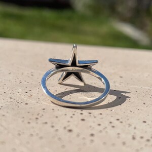 May include: A silver ring featuring a five-pointed star design. The star has a dark interior and is set on a thin, polished band. The ring is shown in a close-up shot, highlighting the details of the star and the band.
