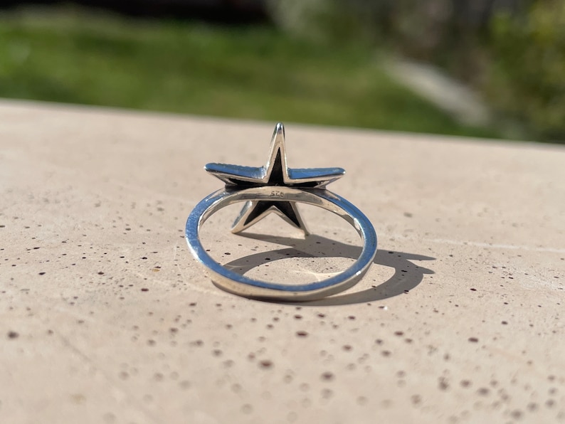 May include: A silver ring featuring a five-pointed star design. The star has a dark interior and is set on a thin, polished band. The ring is shown in a close-up shot, highlighting the details of the star and the band.
