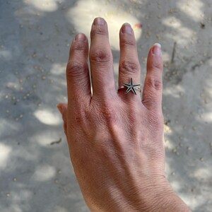 May include: A silver ring featuring a starfish design. The ring is worn on a hand, showcasing the intricate details of the starfish. The ring's design is detailed and eye-catching, perfect for those who love nautical themes.