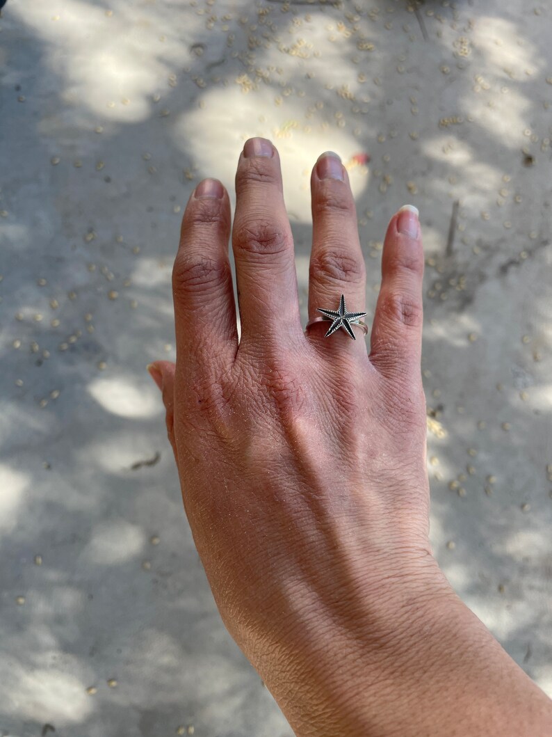 May include: A silver ring featuring a starfish design. The ring is worn on a hand, showcasing the intricate details of the starfish. The ring's design is detailed and eye-catching, perfect for those who love nautical themes.