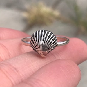 May include: A silver ring with a seashell design. The ring is on a finger.