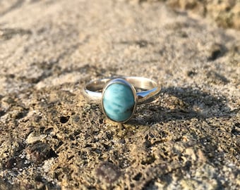May include: A silver ring featuring a blue and white oval gemstone. The ring is displayed on a rough, grey surface.