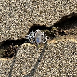 May include: A silver ring with a marquise-shaped, iridescent gemstone. The ring features a Celtic knot design on the sides and a twisted rope border around the stone. The ring is set against a textured, gray surface.