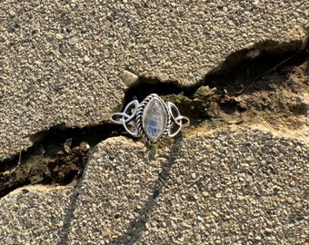 May include: A silver ring with a marquise-shaped, iridescent gemstone. The ring features a Celtic knot design on the sides and a twisted rope border around the stone. The ring is set against a textured, gray surface.