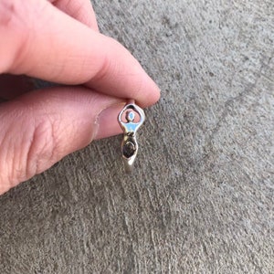 Peut inclure: Une bague en argent repr&eacute;sentant une d&eacute;esse stylis&eacute;e. La bague pr&eacute;sente une t&ecirc;te circulaire, un corps avec un d&eacute;tail en spirale et un petit &eacute;l&eacute;ment circulaire &agrave; la base. La bague est tenue entre les doigts.