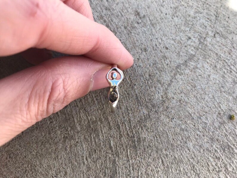Peut inclure: Une bague en argent repr&eacute;sentant une d&eacute;esse stylis&eacute;e. La bague pr&eacute;sente une t&ecirc;te circulaire, un corps avec un d&eacute;tail en spirale et un petit &eacute;l&eacute;ment circulaire &agrave; la base. La bague est tenue entre les doigts.
