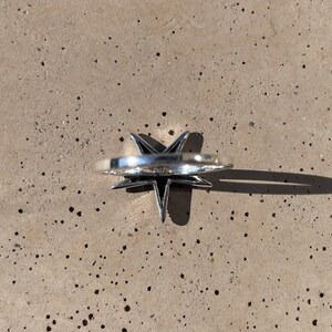 May include: A silver-colored ring with a star-shaped design. The ring's star has pointed edges and a raised center. The ring is set against a speckled, light gray surface, with a shadow cast to the side.