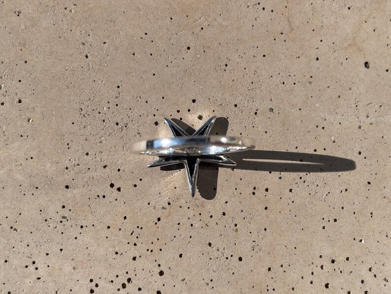 May include: A silver-colored ring with a star-shaped design. The ring's star has pointed edges and a raised center. The ring is set against a speckled, light gray surface, with a shadow cast to the side.